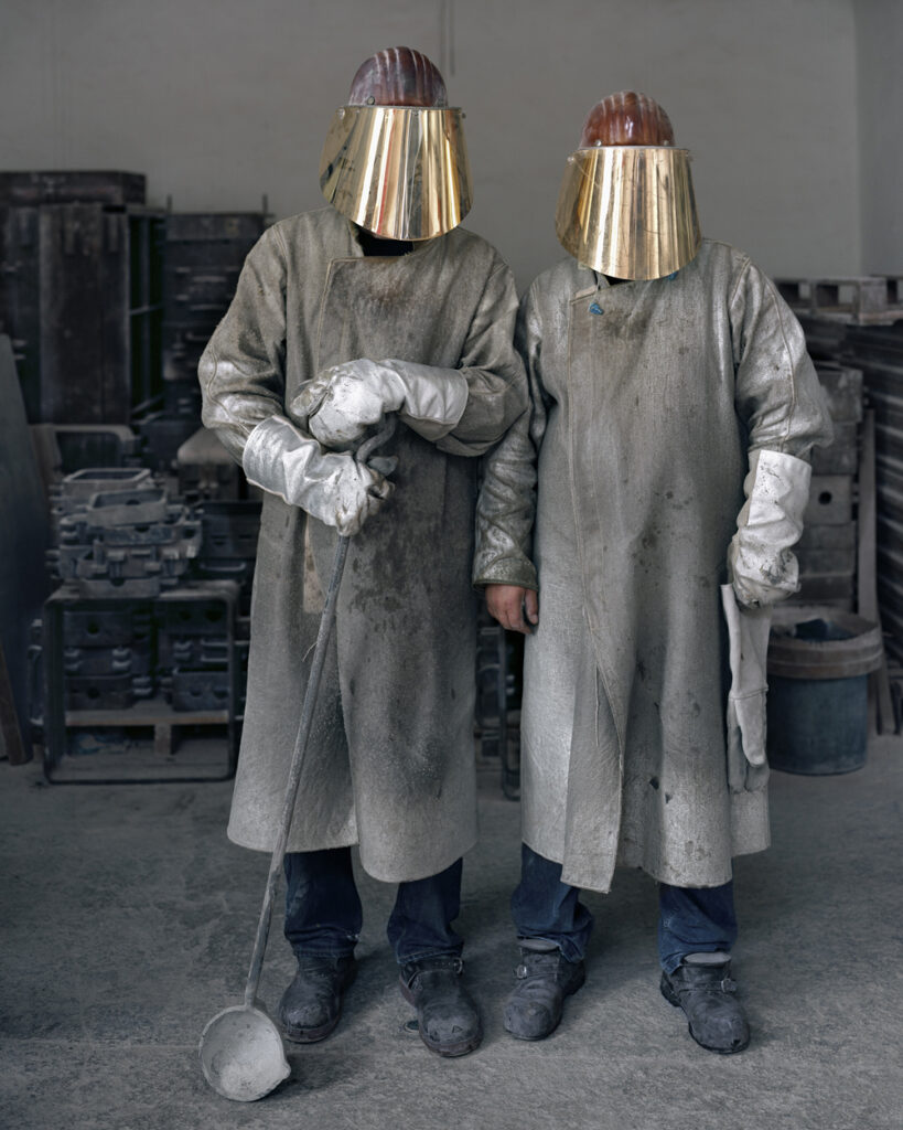Foto von Lukas Einsele: zwei Metallgießer in grauer Schutzkleidung. Ihre Gesichter sind von einem goldenem Visier verdeckt