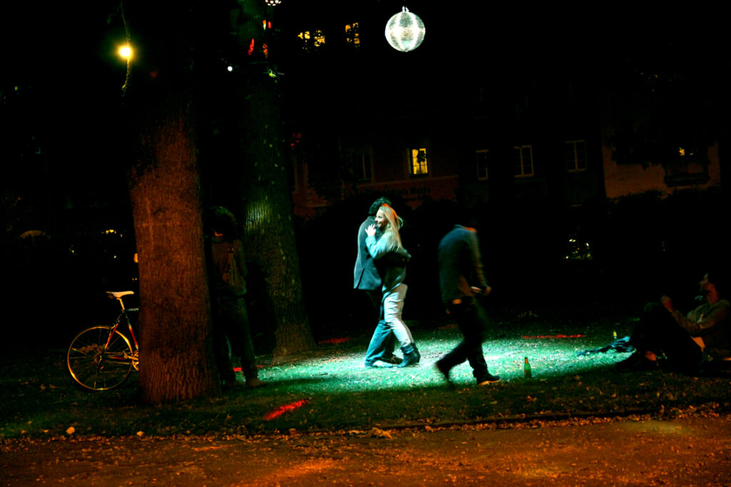 Ein Paar tanzt auf einer beleuchteten Tanzfläche. Diese befindet sich draussen, im öffentlichen Raum. Es ist Nacht und über ihnen schwebt eine Discokugel